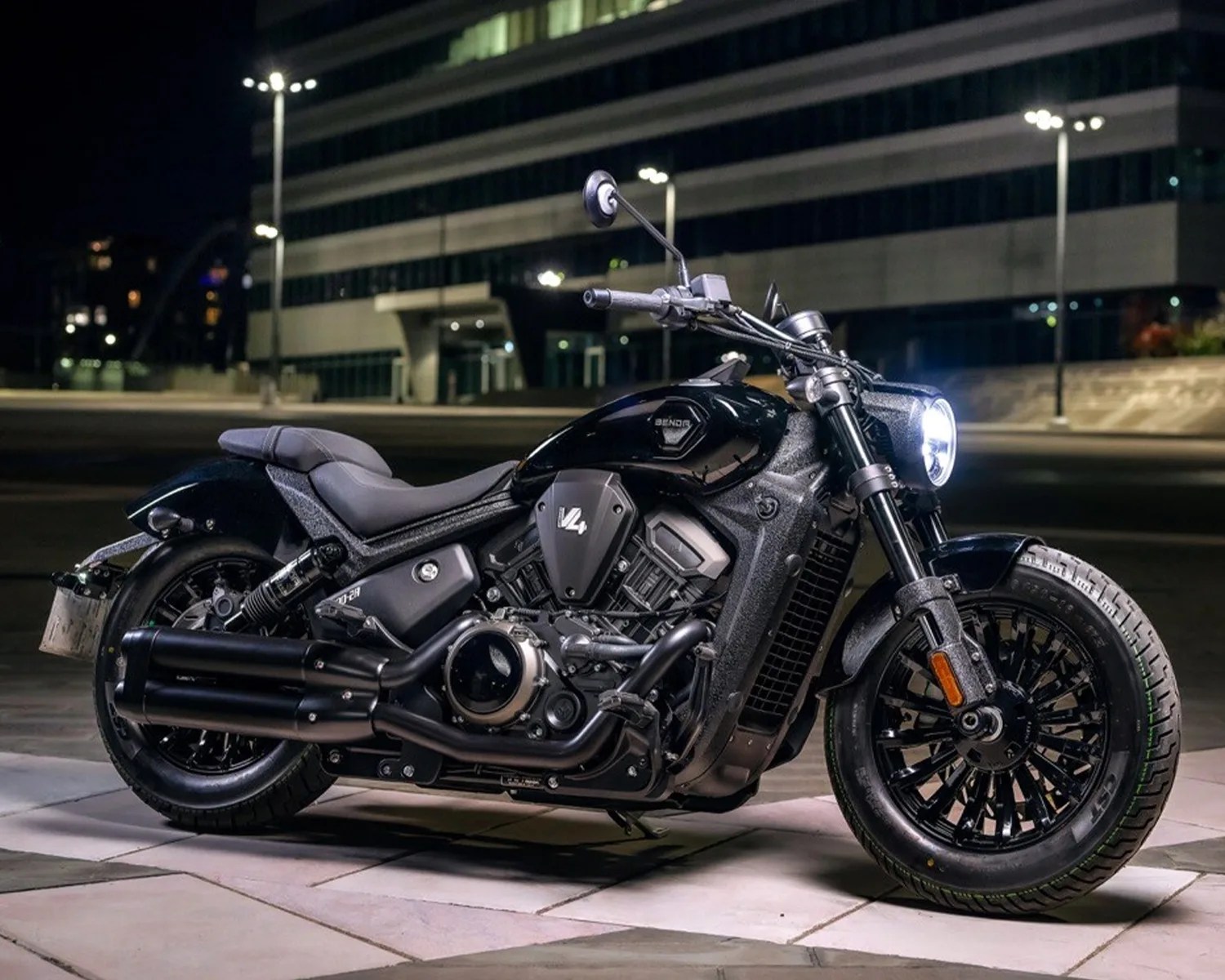 Black cruiser motorcycle with V4 engine and dual exhaust pipes parked on tiled pavement at night.