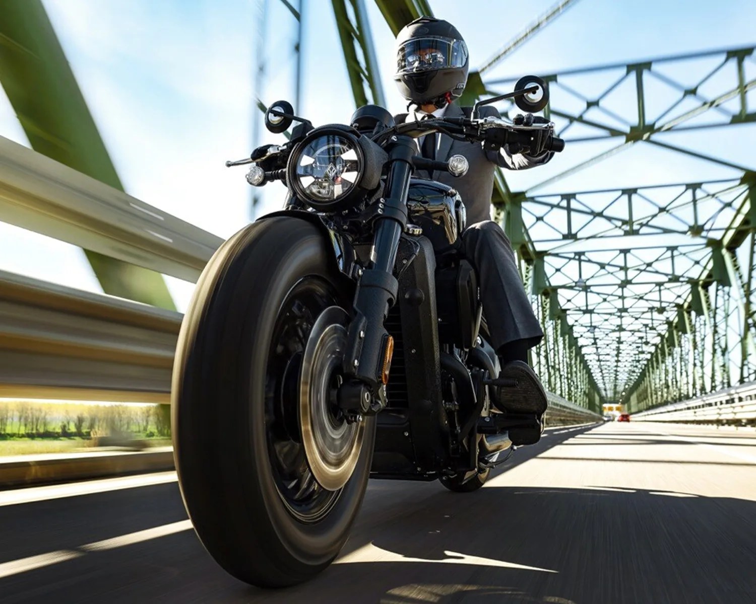 Black motorcycle with round headlight ridden by a person wearing a black helmet on a green metal bridge.