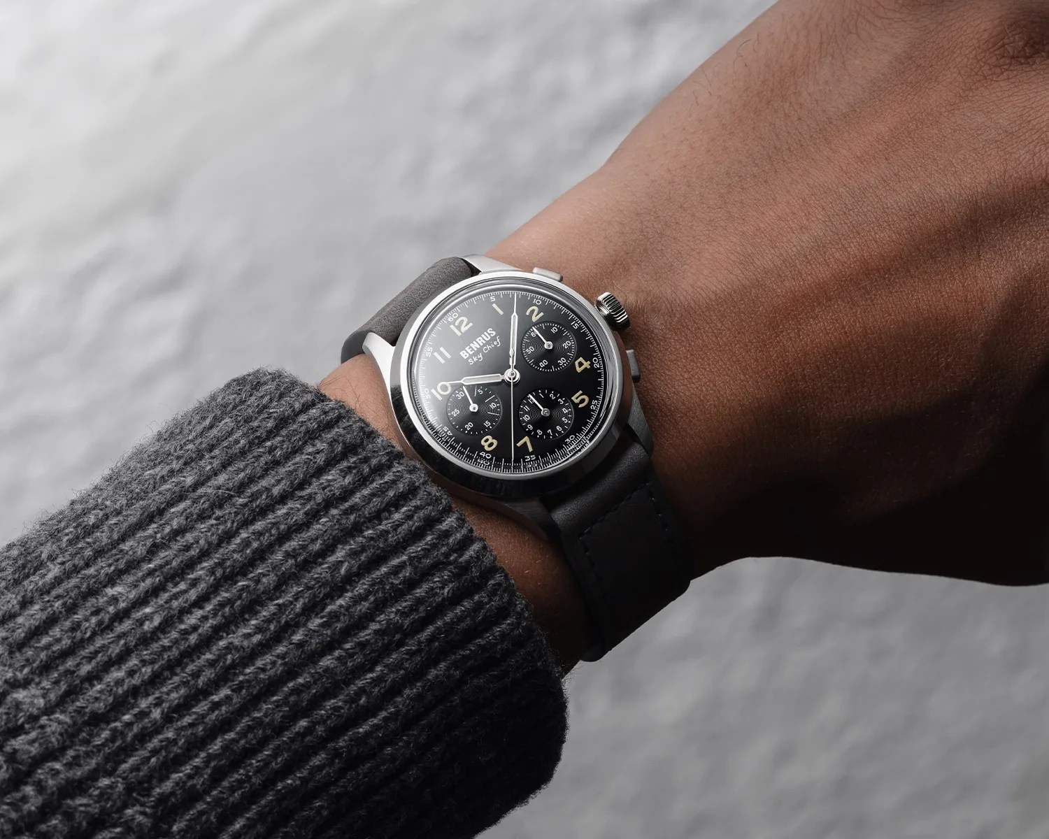 Wristwatch with black dial, three subdials, and gray leather strap worn on a wrist with a gray knitted sleeve.