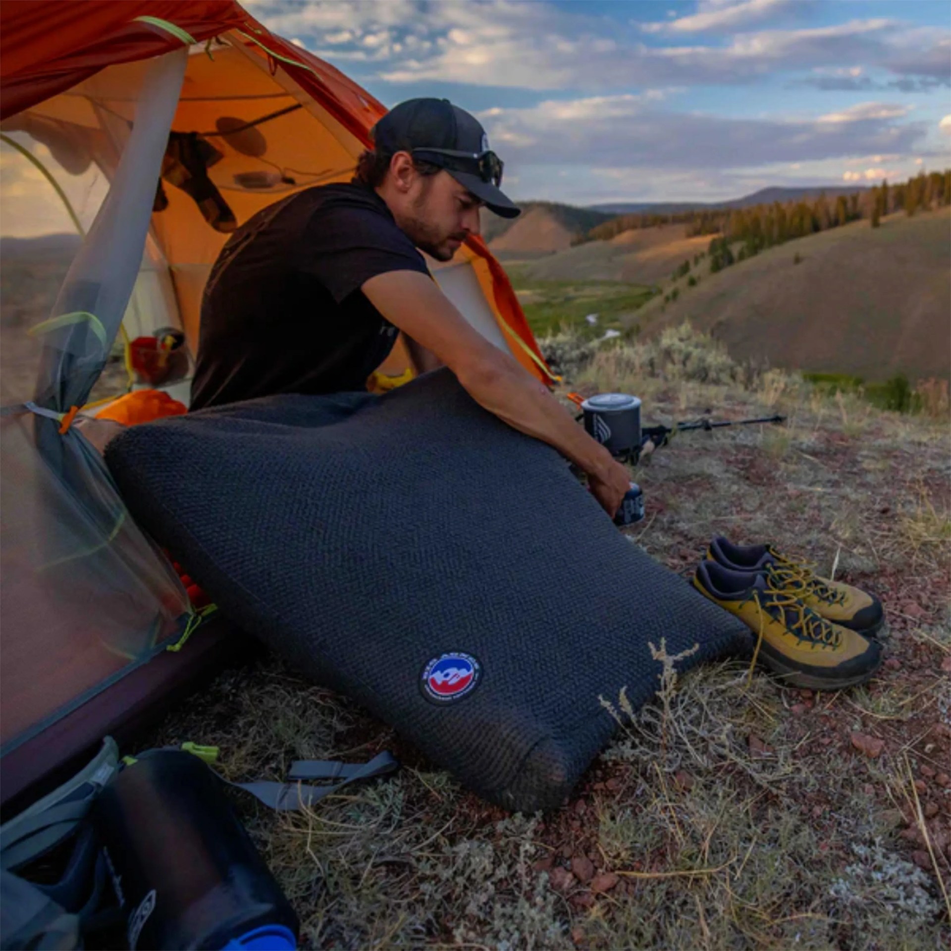 Man sitting in an orange tent holding a large dark gray camping pillow with hiking shoes and gear outside.