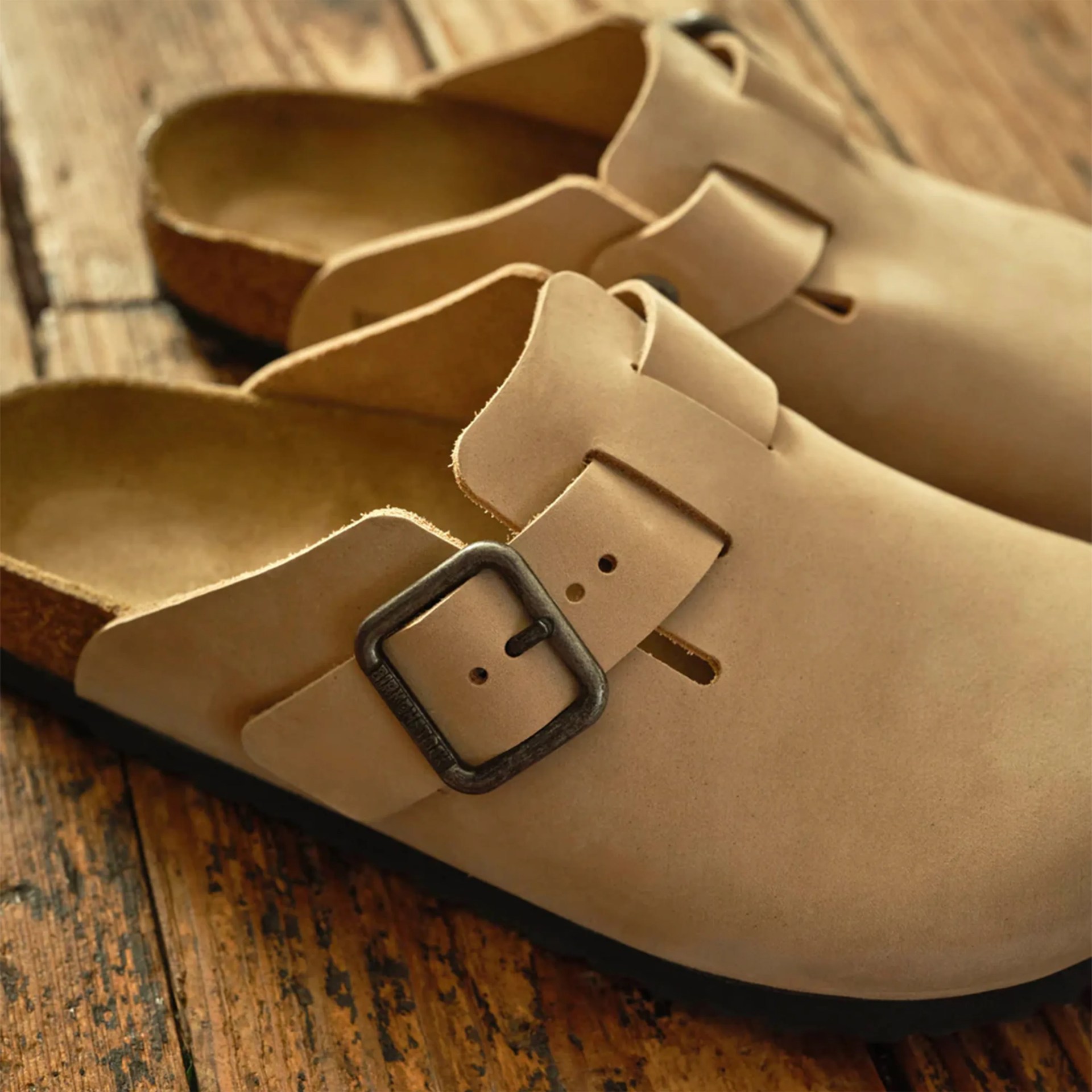 Pair of beige leather slip-on clogs with black metal buckles on a wooden floor.