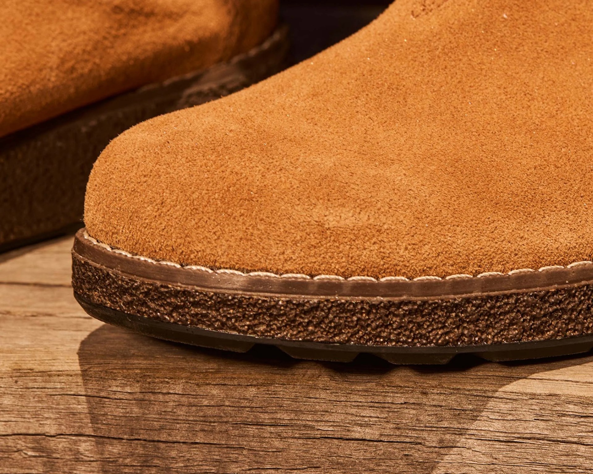 Close-up of a tan suede shoe toe with textured dark brown sole and white stitching on wooden surface.