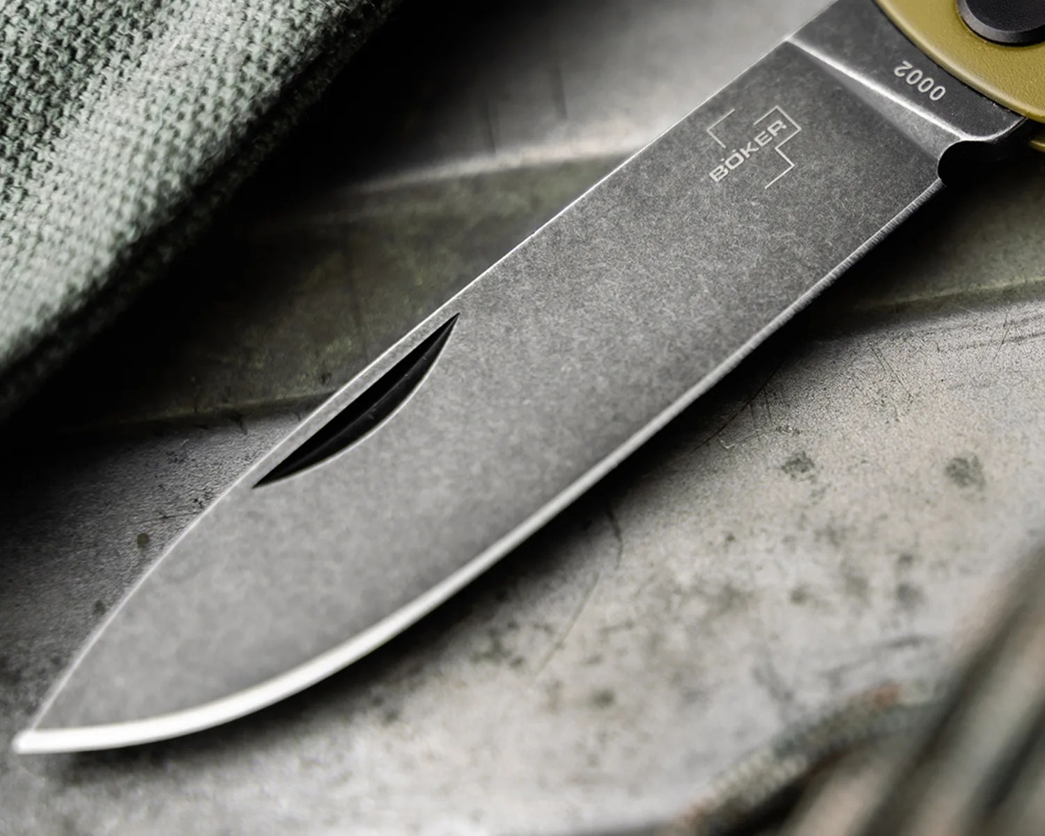 Close-up of a stonewashed B&ouml;ker folding knife blade with a nail nick on a textured surface.