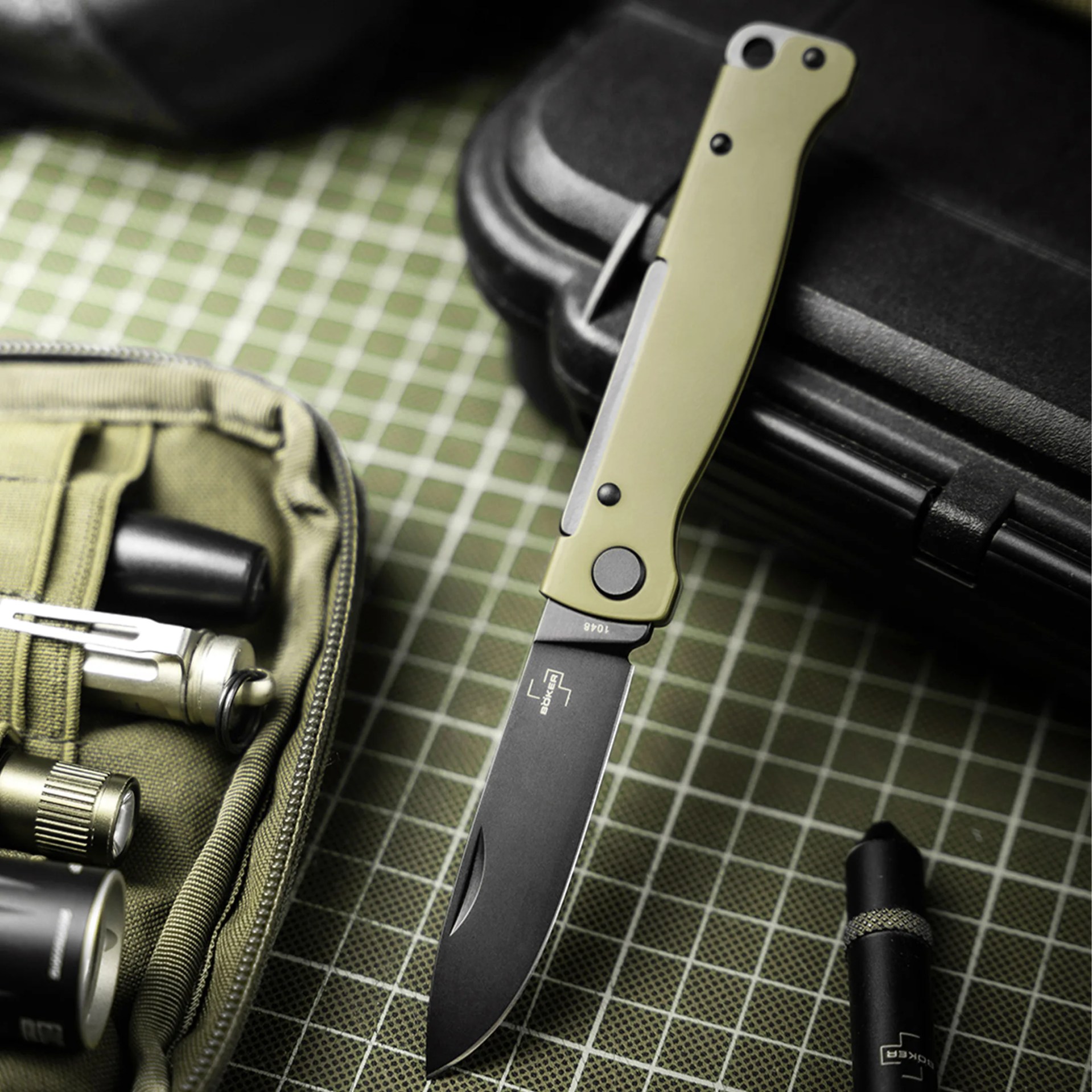 Folding knife with olive green handle and black blade labeled "B&ouml;ker" on a green grid surface next to a black case and green pouch.