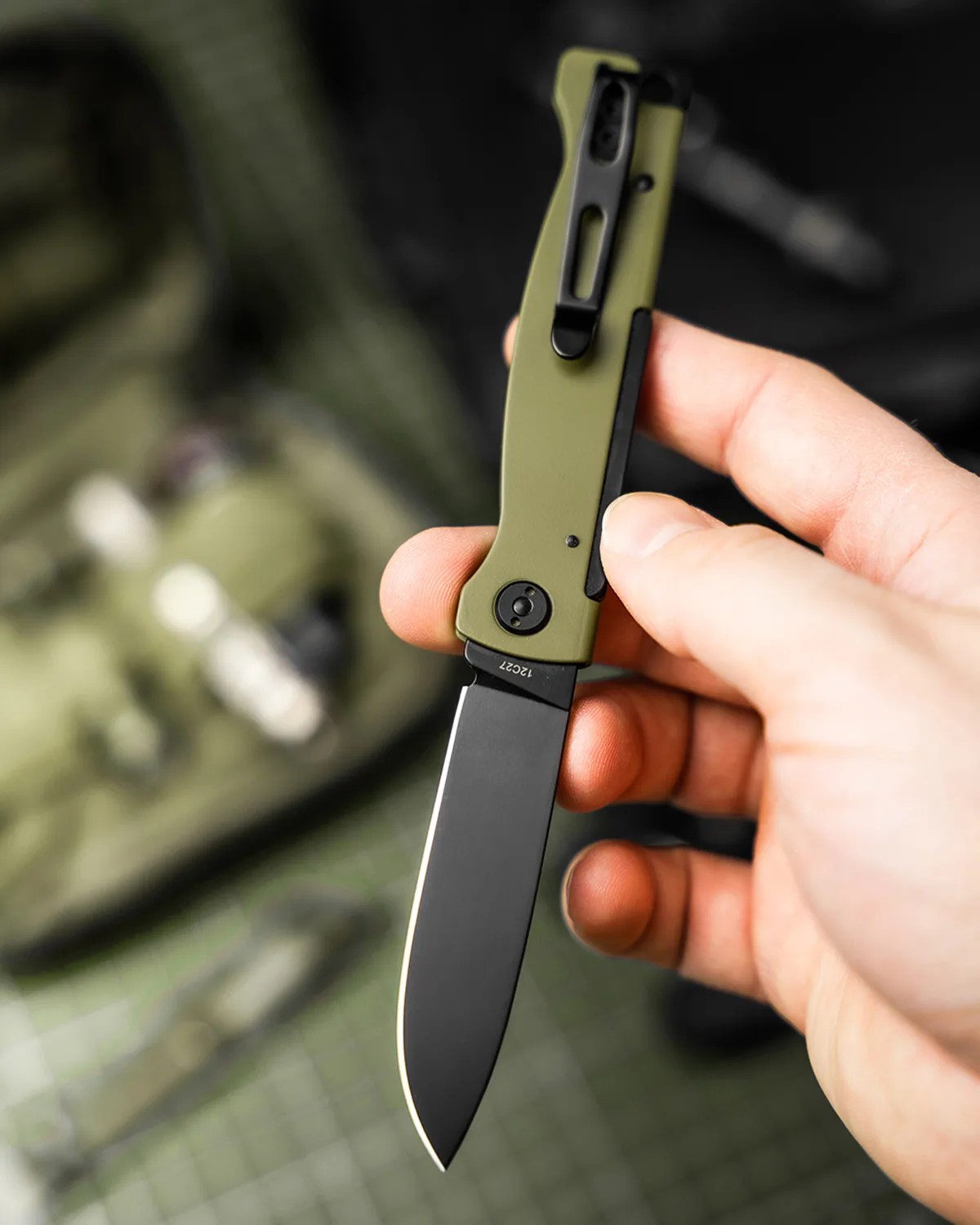 Hand holding a folding knife with an olive green handle and black blade.