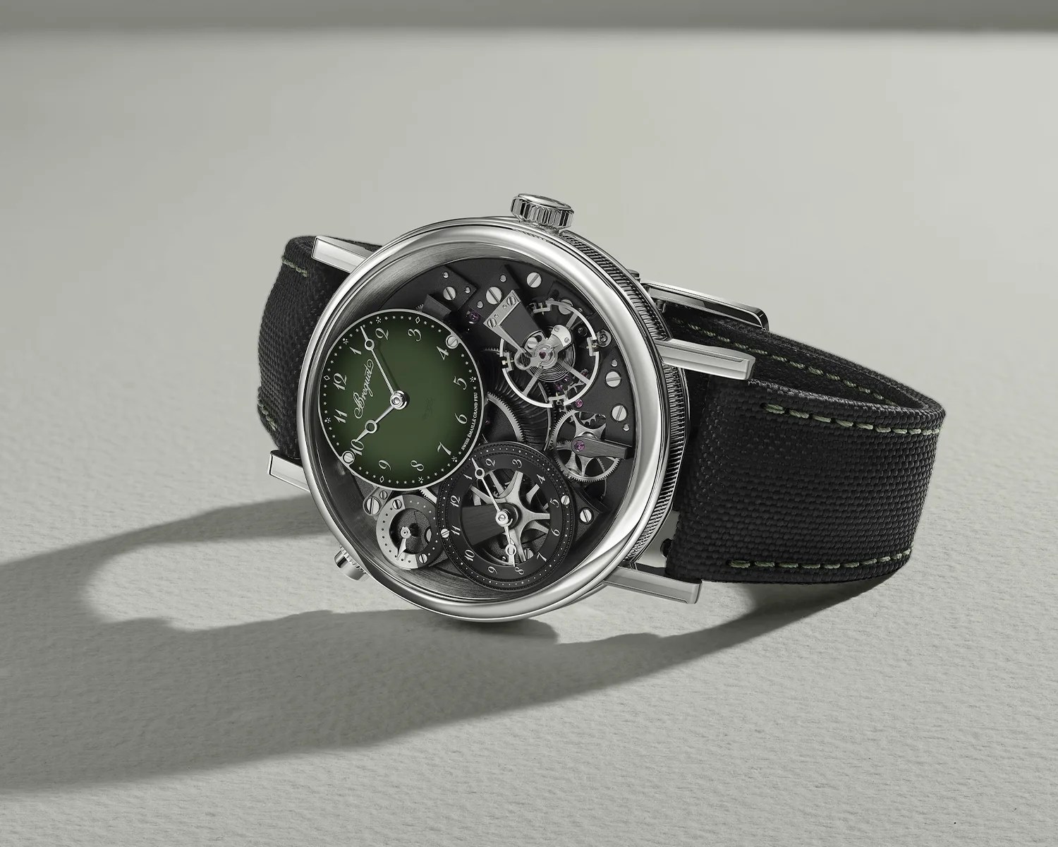 Silver mechanical wristwatch with exposed gears, green subdial, and black fabric strap with green stitching.