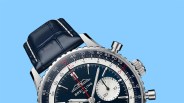 Close-up of a Breitling watch with a blue dial, silver subdial, and dark blue leather strap against a blue background.