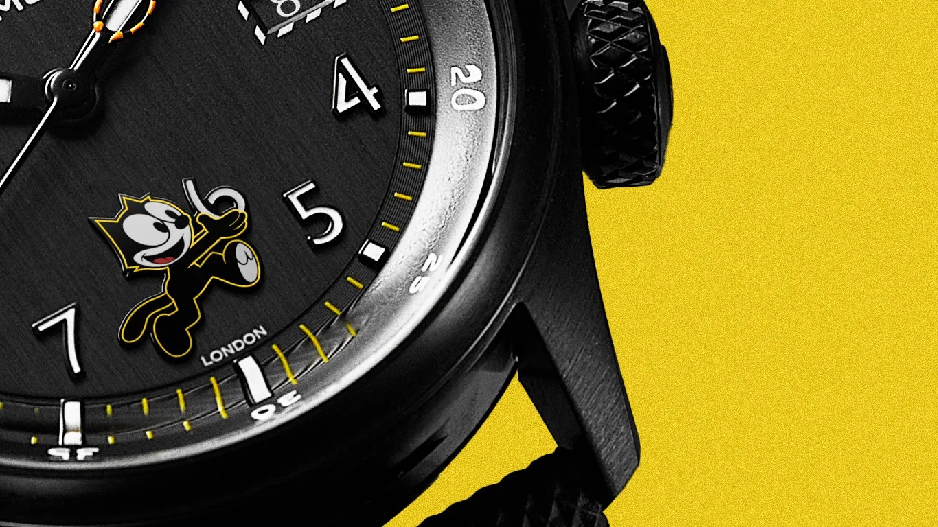 Close-up of a black watch face with white numerals, yellow minute markers, and a small Felix the Cat figure above the word "London."