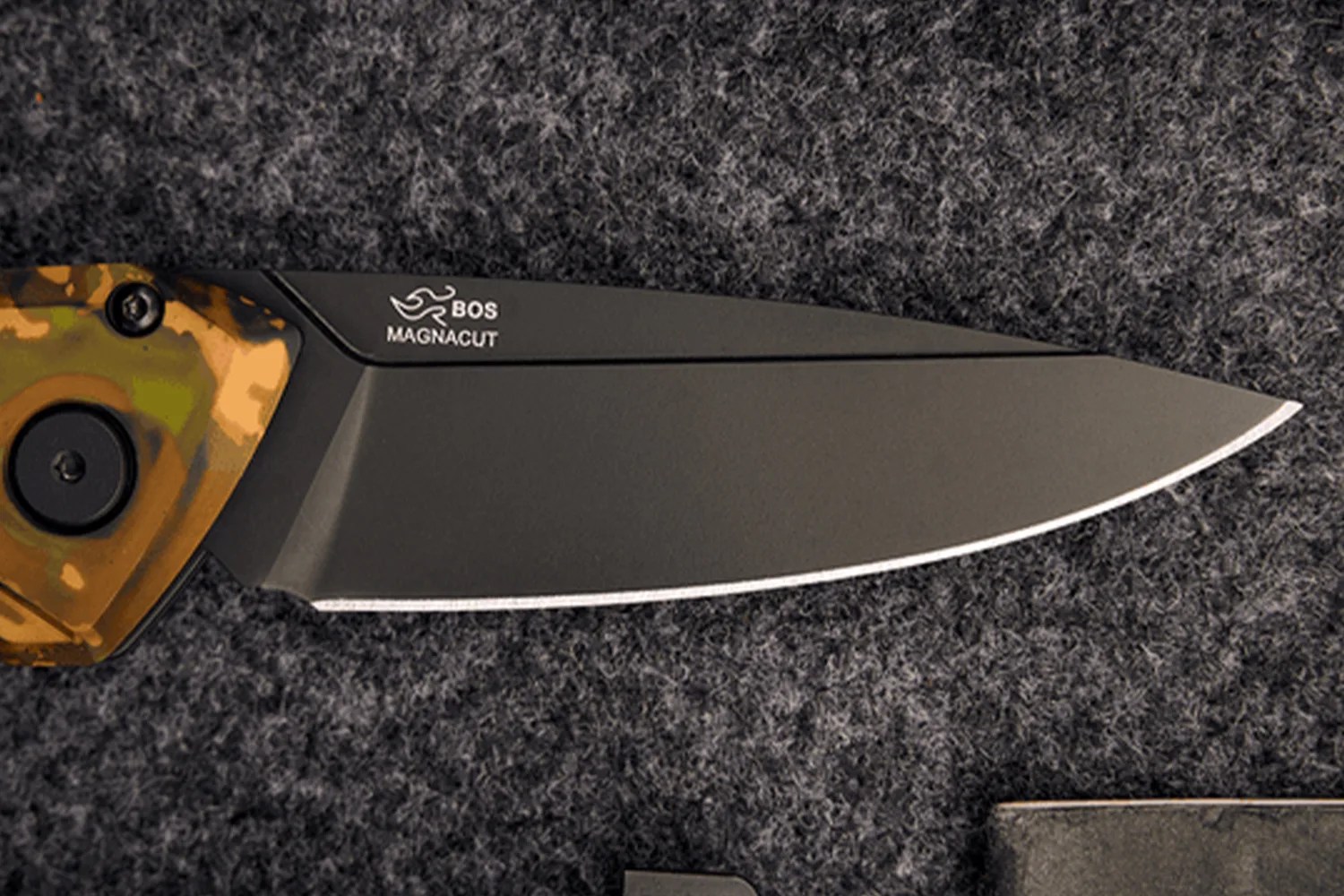Black BOS Magnacut folding knife blade with a camo handle on a dark textured surface.