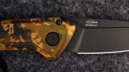 Black folding knife blade with "BOS MAGNACUT" logo and camo-patterned handle on a dark textured surface.