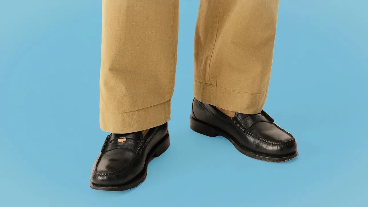 Black leather loafers with a small gold emblem worn with tan trousers against a blue background.