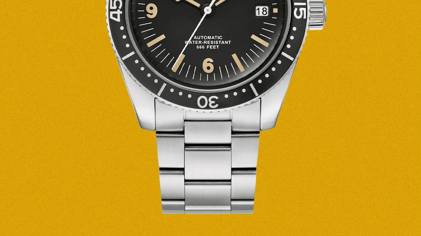 Close-up of a stainless steel watch bracelet attached to a black dive watch with a rotating bezel on a mustard yellow background.