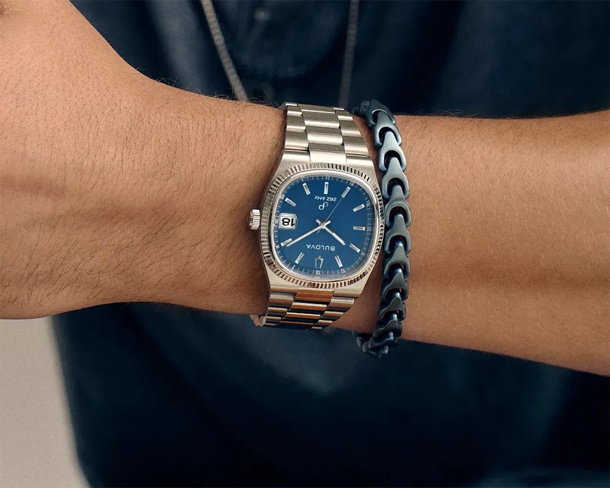 Silver metal Bulova wristwatch with a blue dial and date display worn on a wrist next to a black chain bracelet.