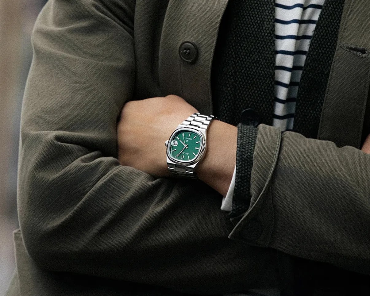 Silver metal wristwatch with a green square dial worn on a wrist crossed over an arm in an olive green jacket.