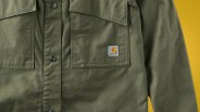 Olive green Carhartt jacket with snap buttons and chest pockets, shown against a yellow background.