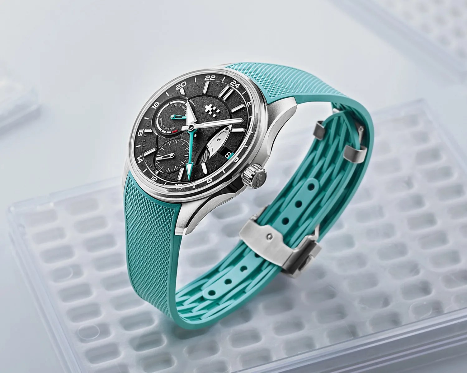 Silver wristwatch with black textured dial and turquoise rubber strap on a clear plastic surface.