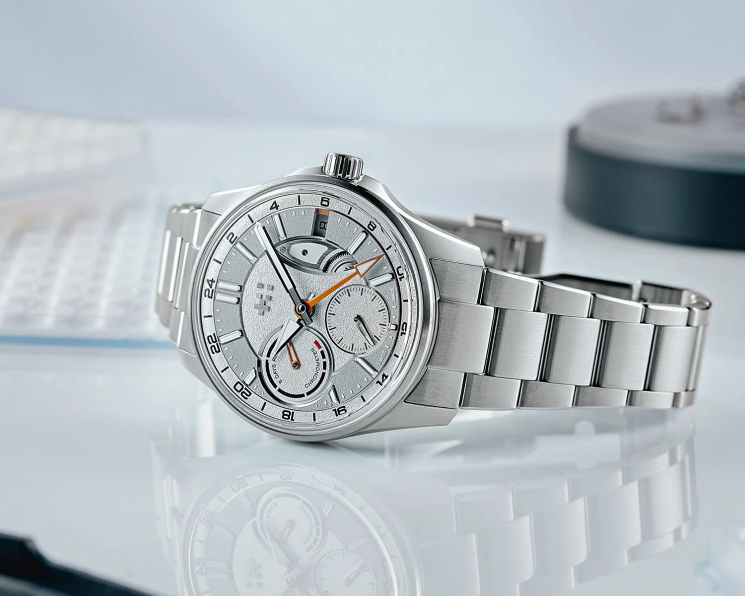 Silver stainless steel wristwatch with textured dial, orange accents, and metal link bracelet on reflective surface.