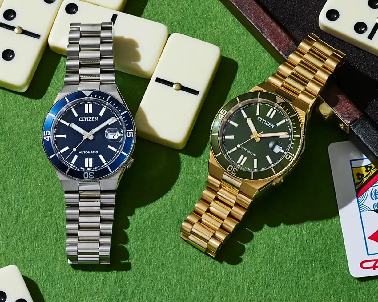 Two Citizen automatic wristwatches, one silver with a blue dial and one gold with a black dial, on a green felt surface with dominoes and a playing card.