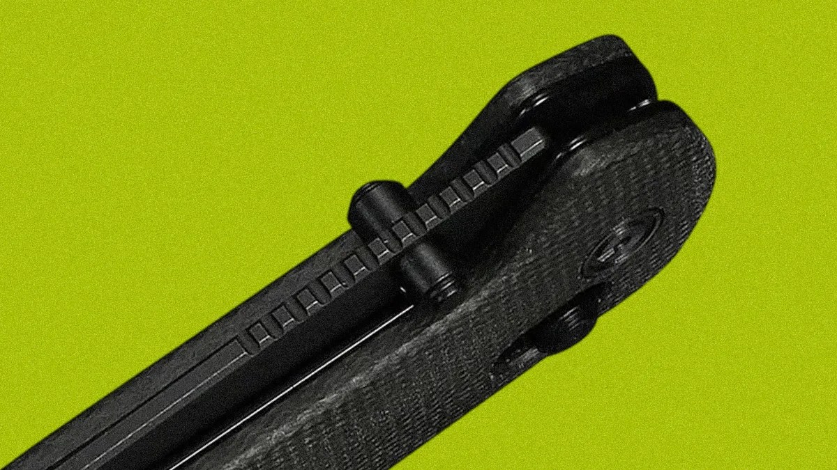 Close-up of a black folding knife hinge and textured handle against a green background.