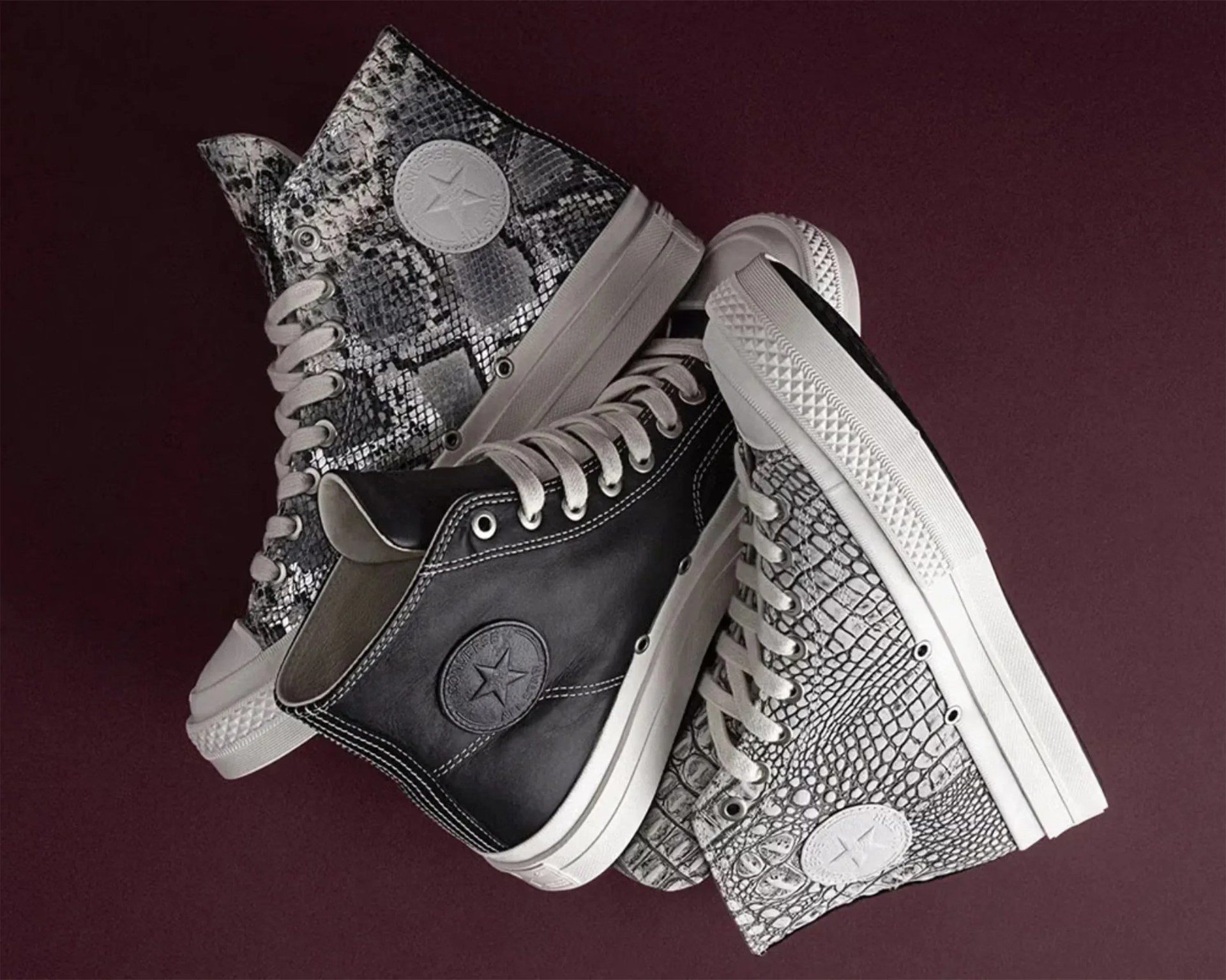 Three pairs of high-top Converse sneakers in black leather, black and white snakeskin, and white crocodile pattern on a dark purple background.