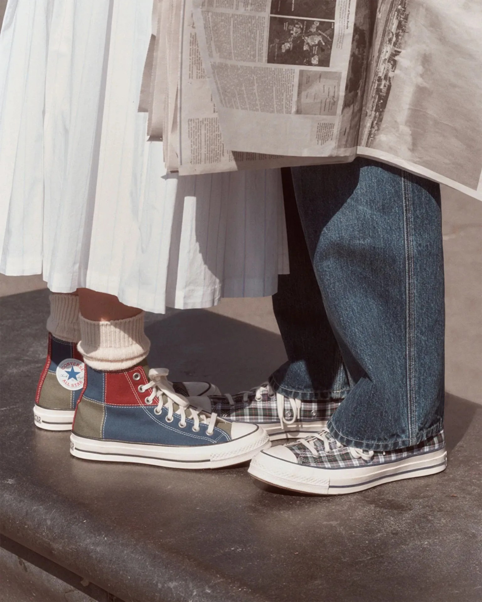 Pair of feet wearing colorful patchwork Converse high-top sneakers and plaid low-top sneakers, with a white pleated skirt and blue jeans visible.