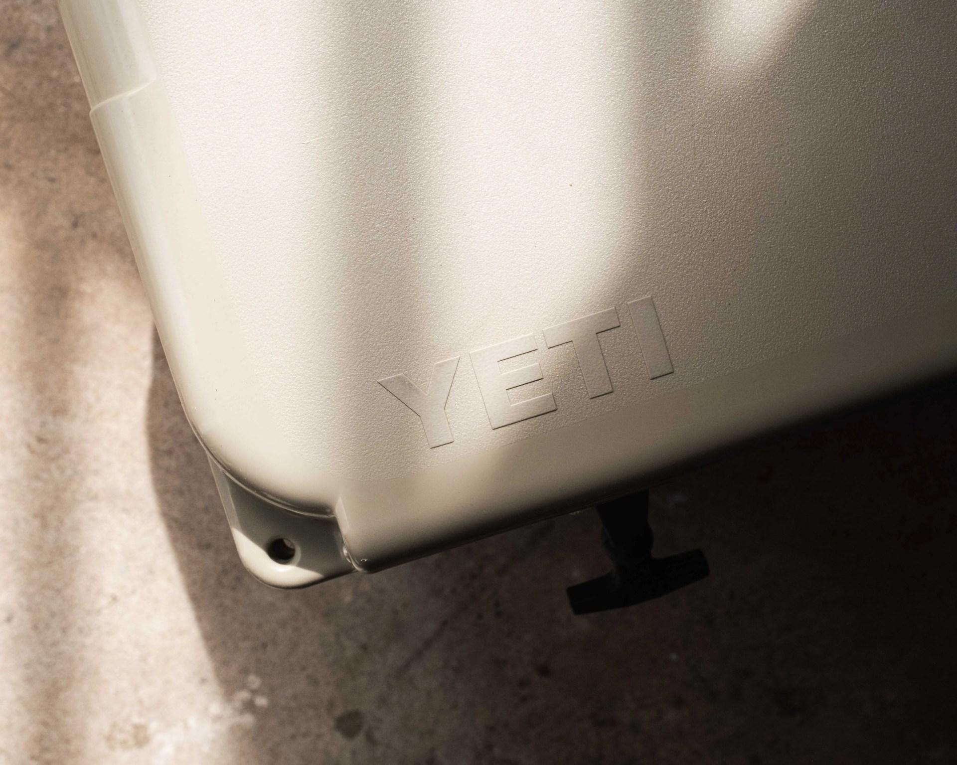 Close-up of a beige YETI cooler corner with embossed logo and shadow on a concrete floor.