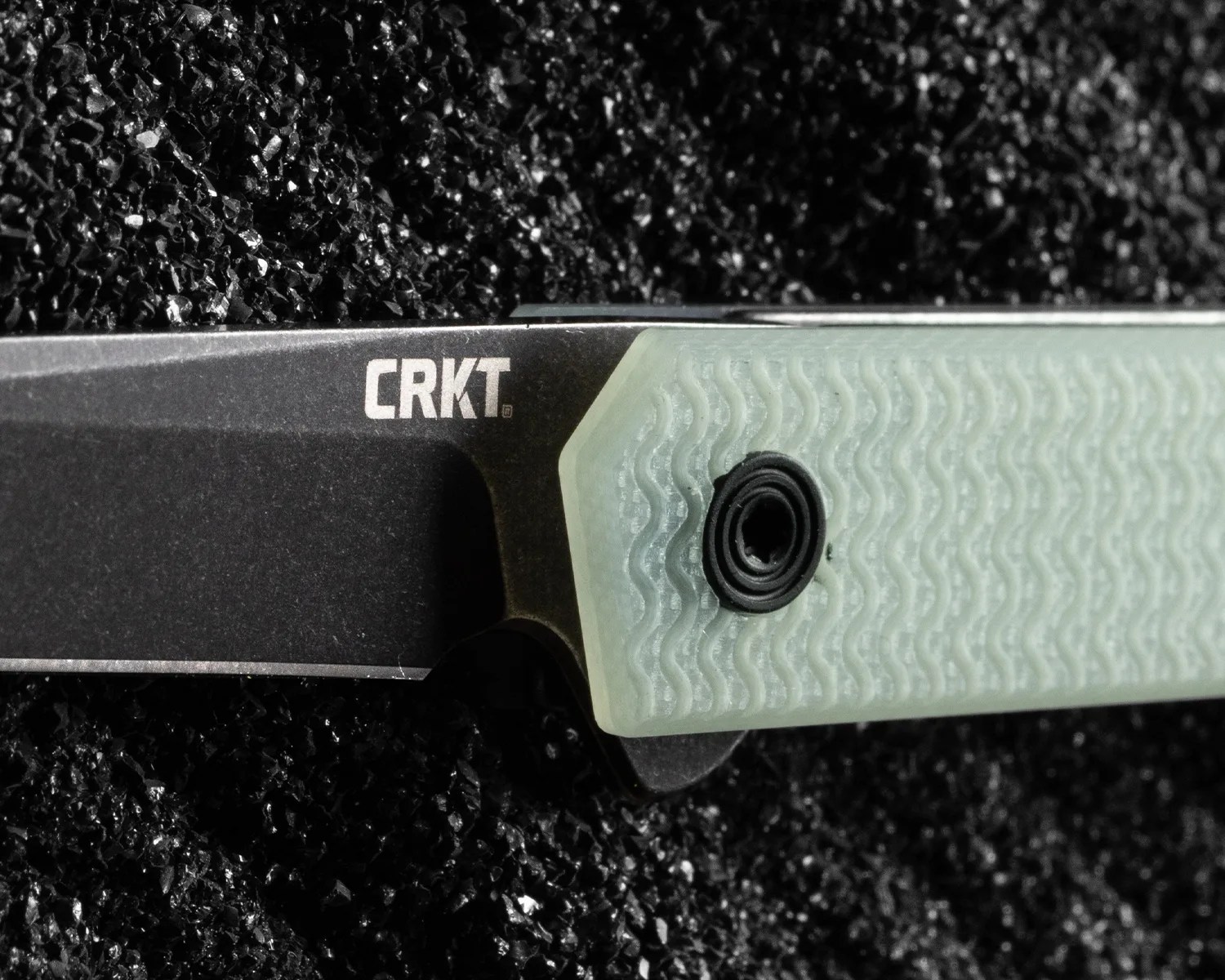 Close-up of a black CRKT knife blade with a textured light green handle on a black granular surface.