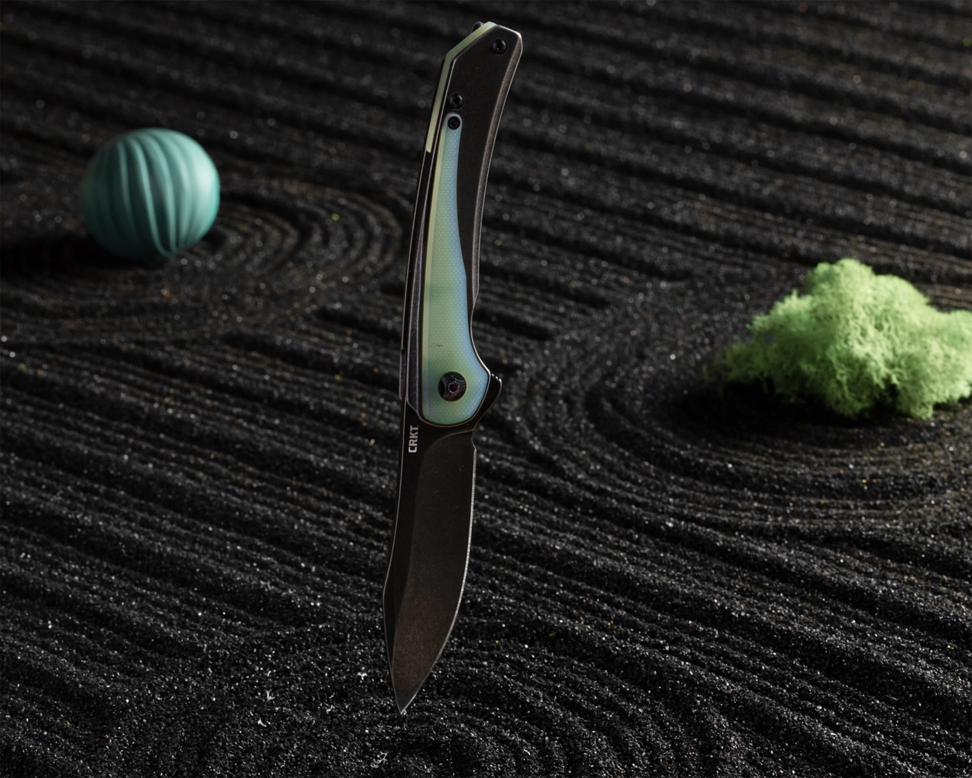 Black folding knife with a translucent light blue handle standing upright on textured black sand with a green moss and blue ball in the background.