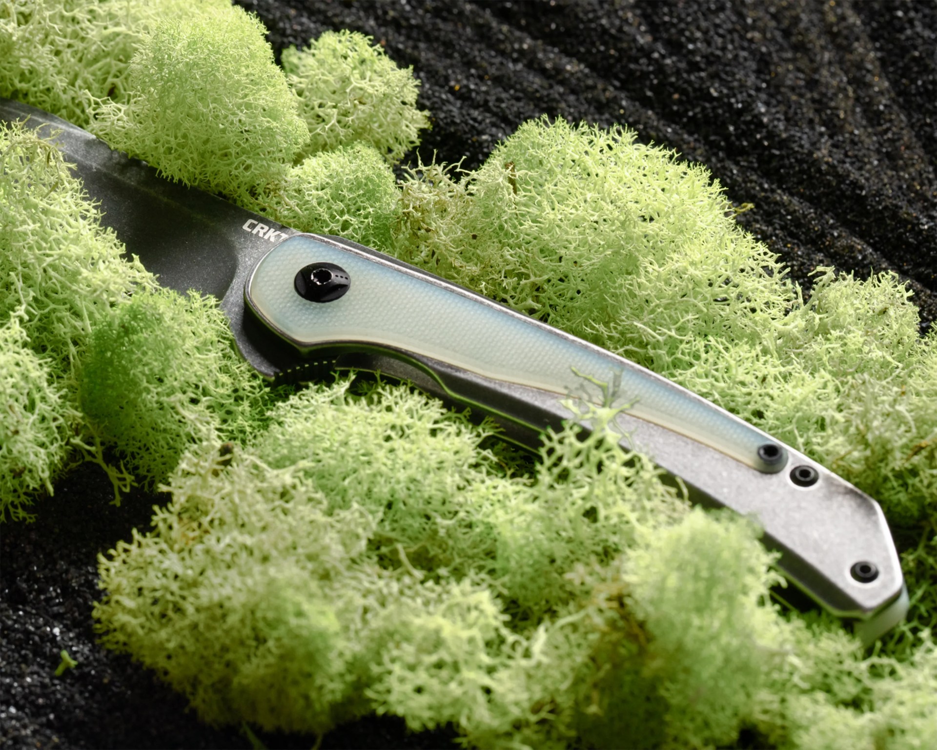 Folding knife with a light blue textured handle and black blade partially covered by green moss on dark soil.