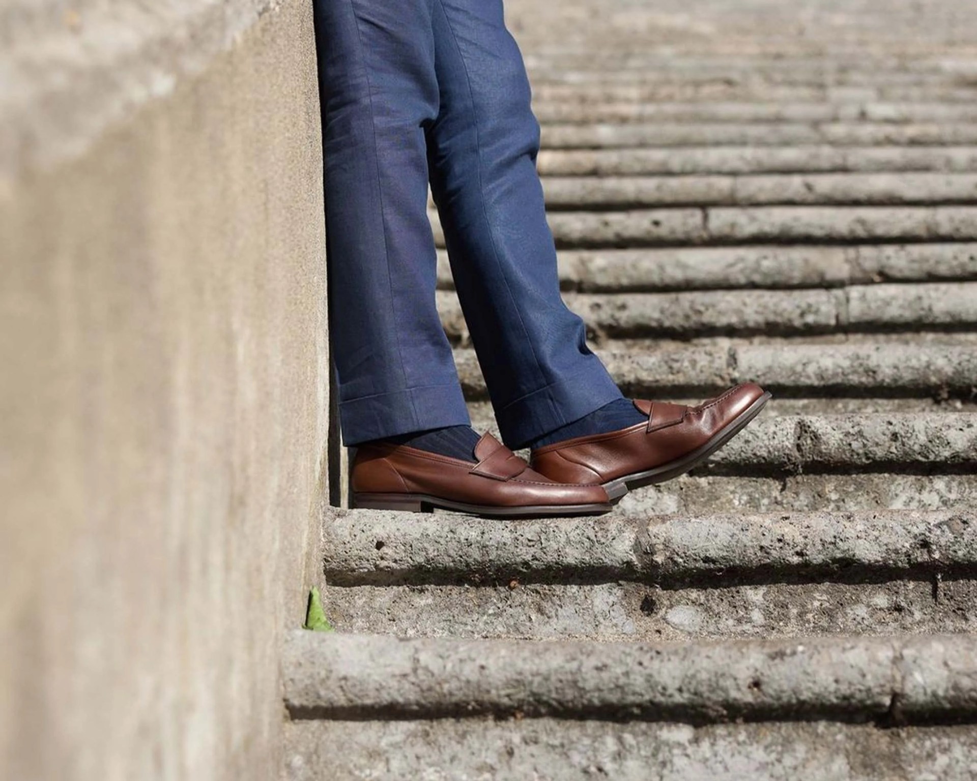 Brown leather loafers worn with navy blue pants on stone steps.