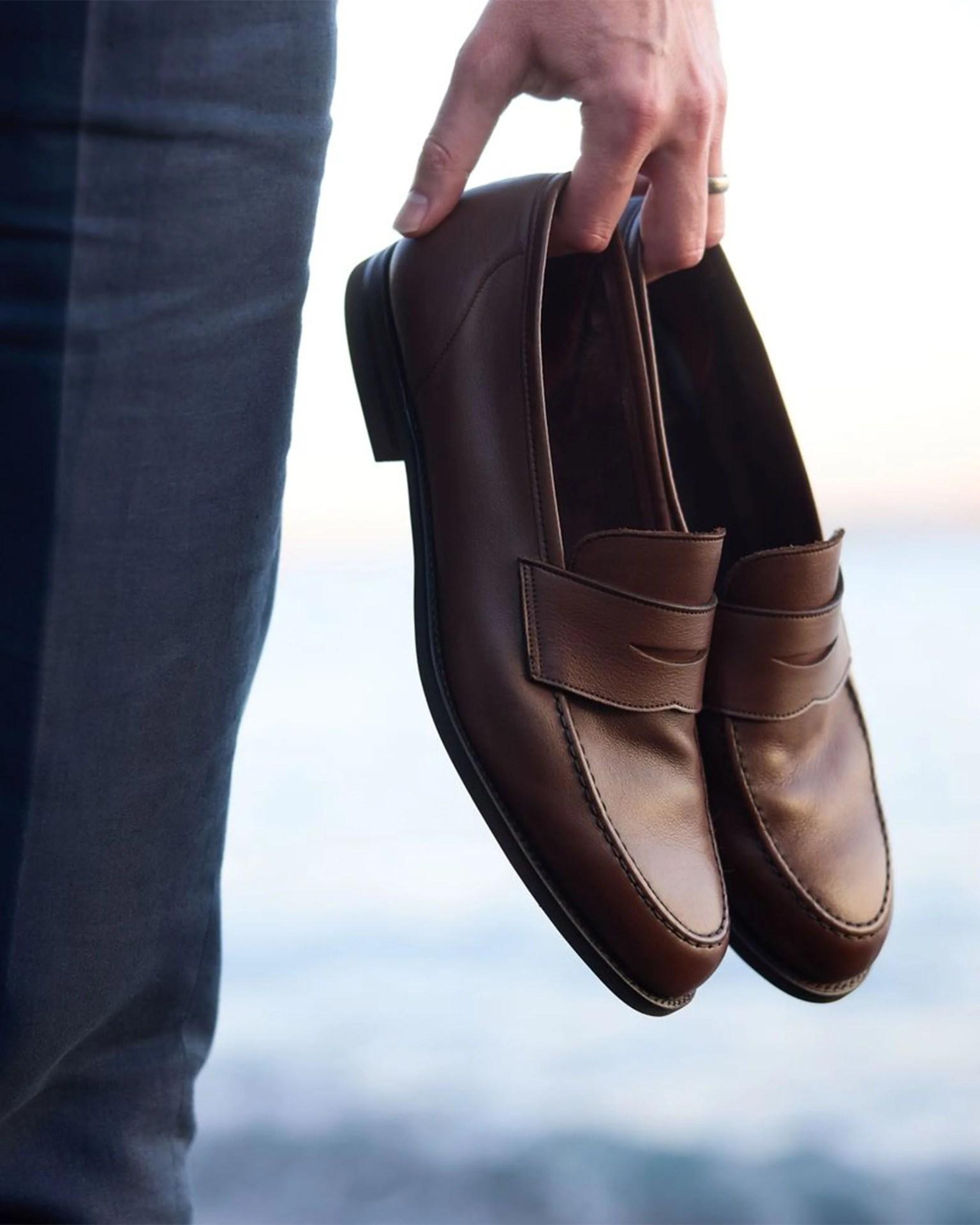 Hand holding a pair of brown leather loafers with black soles near the ocean.