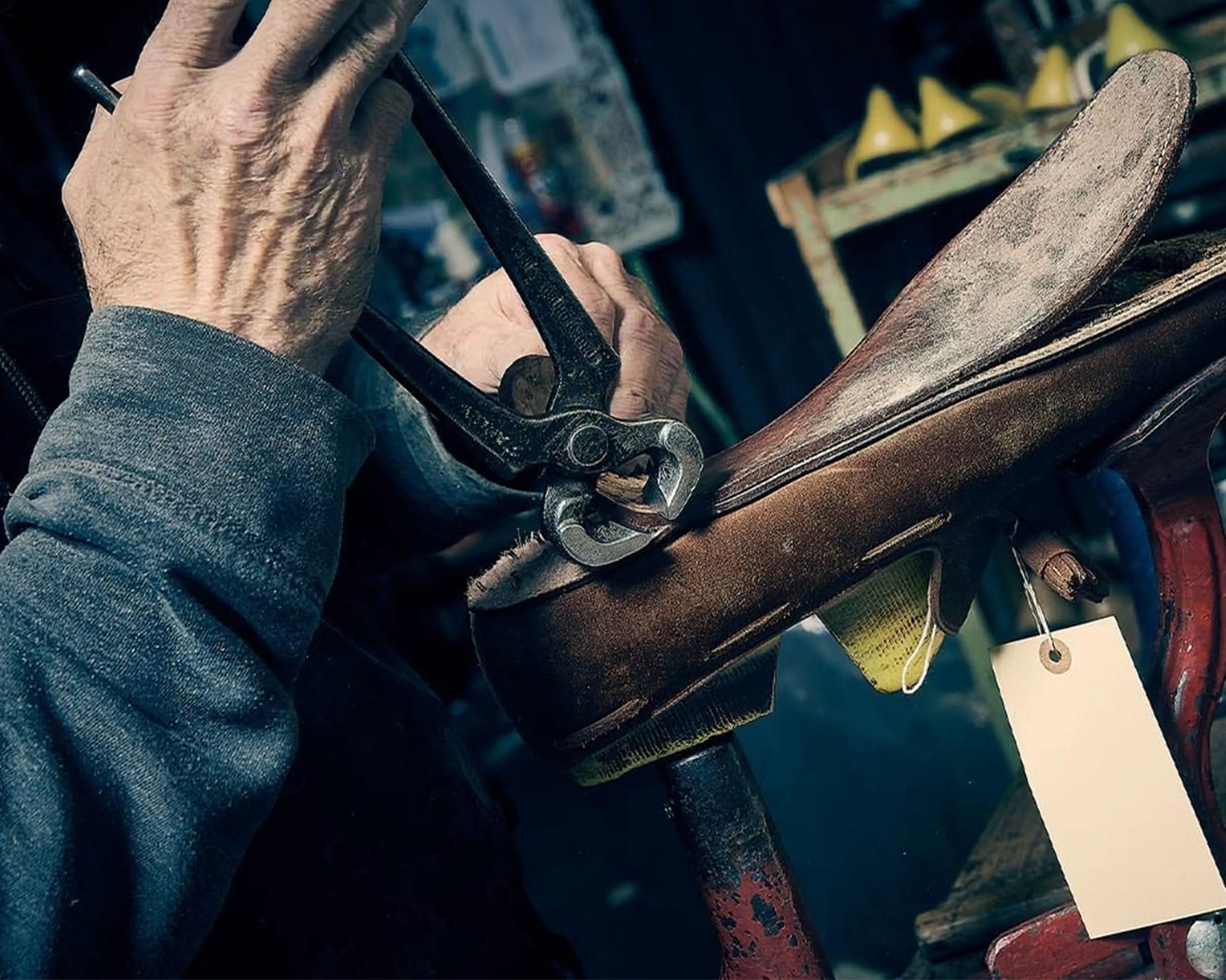 Hands using pliers to repair a brown leather shoe on a shoe stand with a blank tag attached.