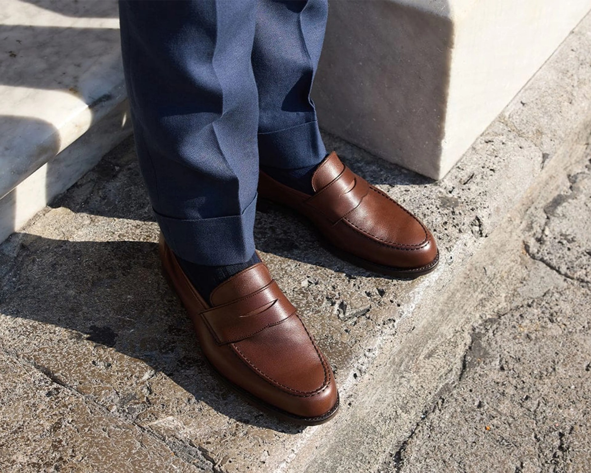 Brown leather penny loafers worn with navy trousers on stone steps.
