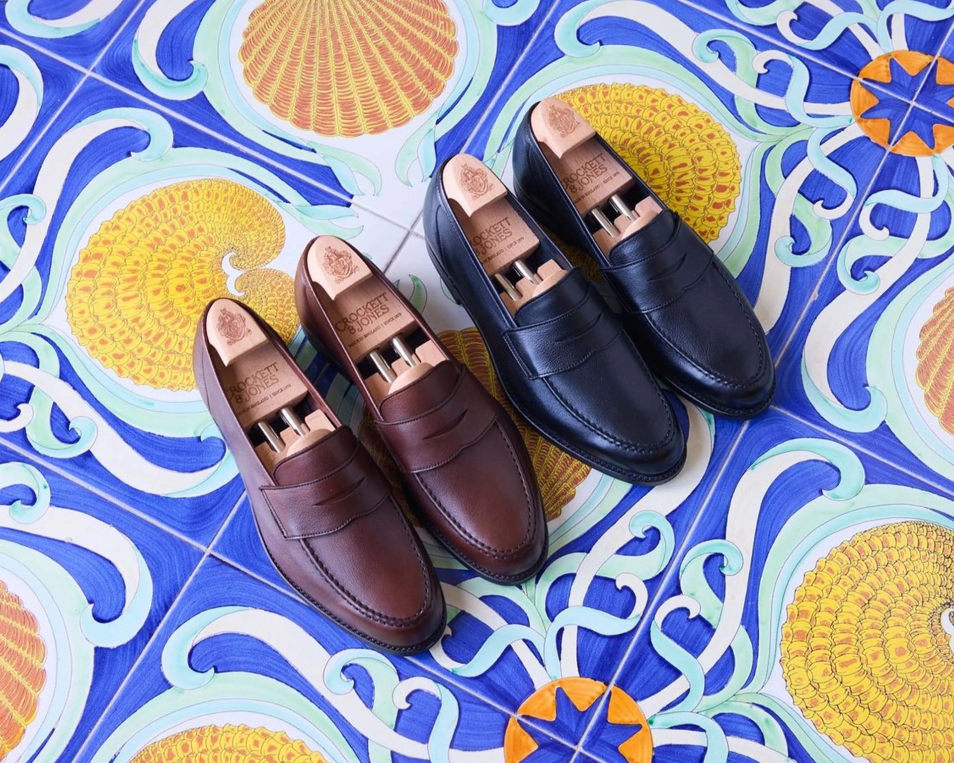 Two pairs of leather loafers, one brown and one black, each with wooden shoe trees inside, placed on colorful patterned tiles.