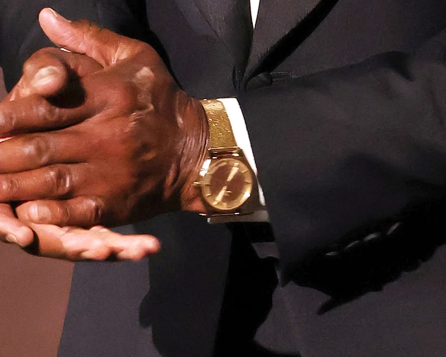 Gold wristwatch with a textured band worn under a dark suit jacket on a person's wrist.
