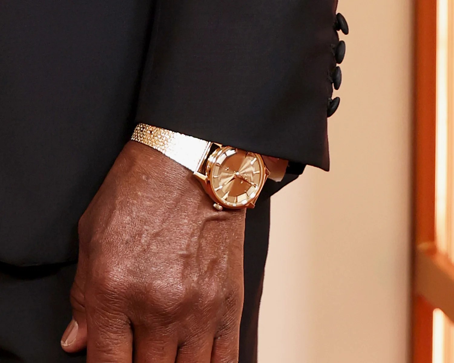 Gold wristwatch with a textured metal band worn under a black suit sleeve.