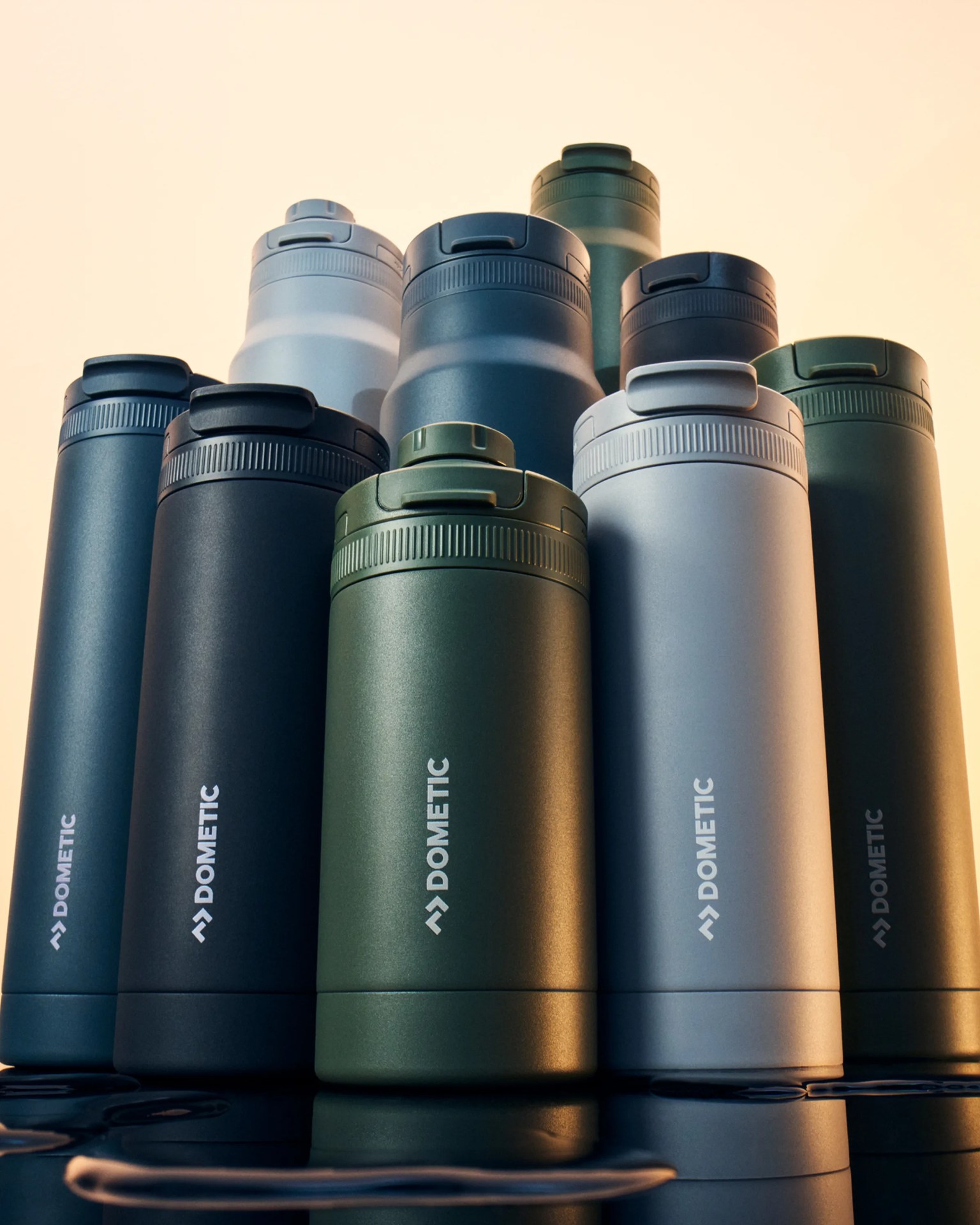 Ten matte-finish Dometic insulated bottles in green, black, blue, and gray arranged in a pyramid on a reflective surface.