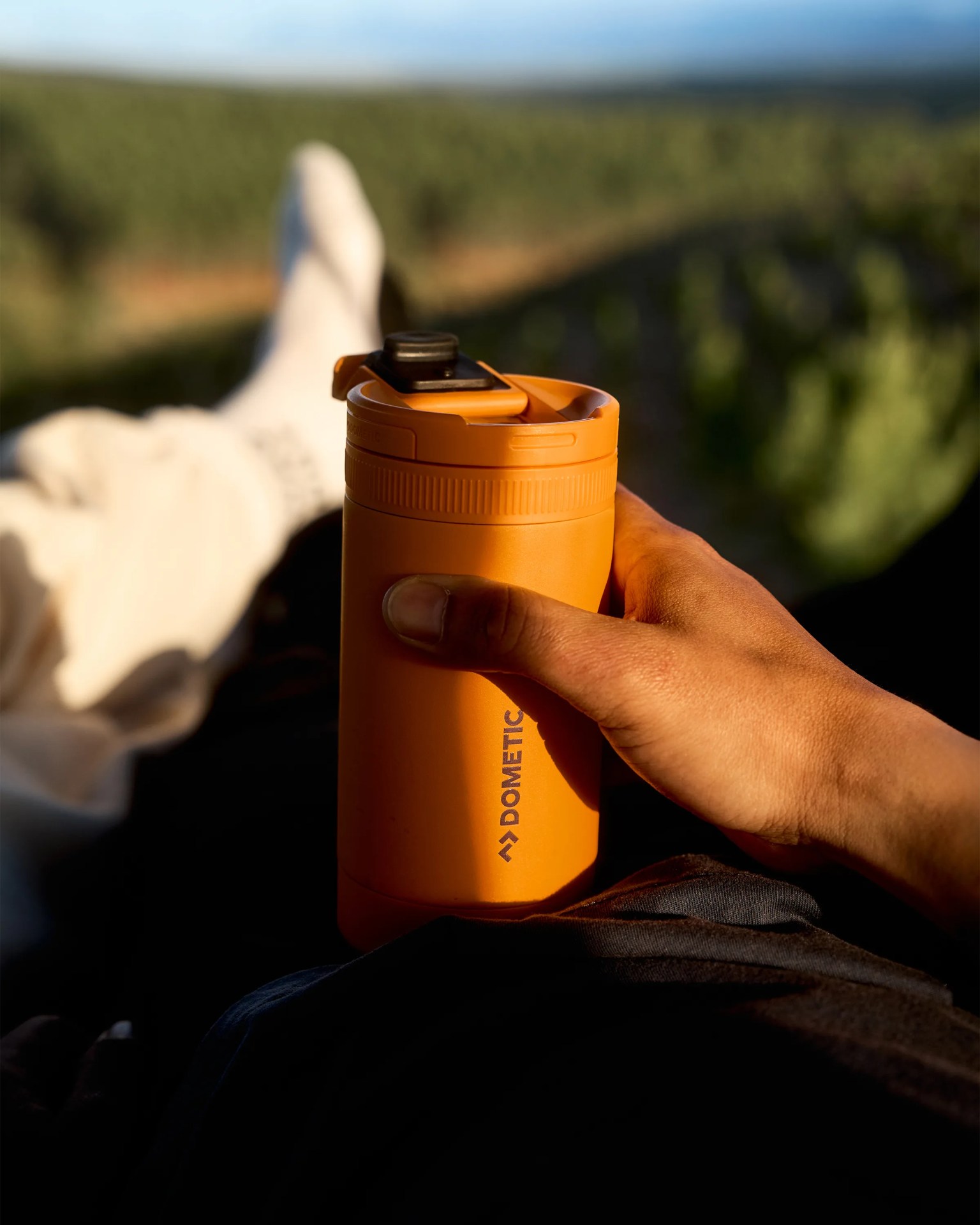 Orange Dometic insulated travel mug held in a hand with blurred outdoor background.