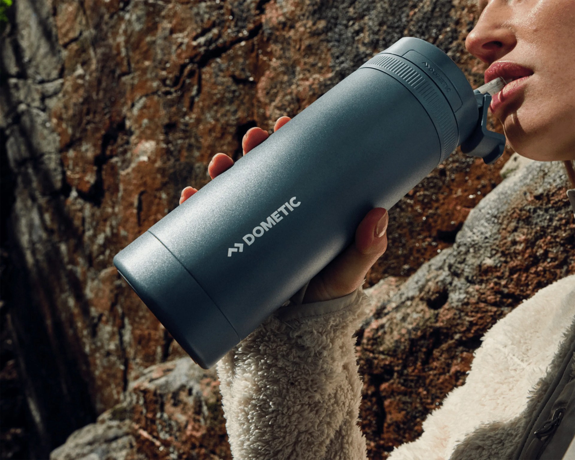 Blue Dometic insulated water bottle with flip-top straw held by a person wearing a beige fleece jacket.