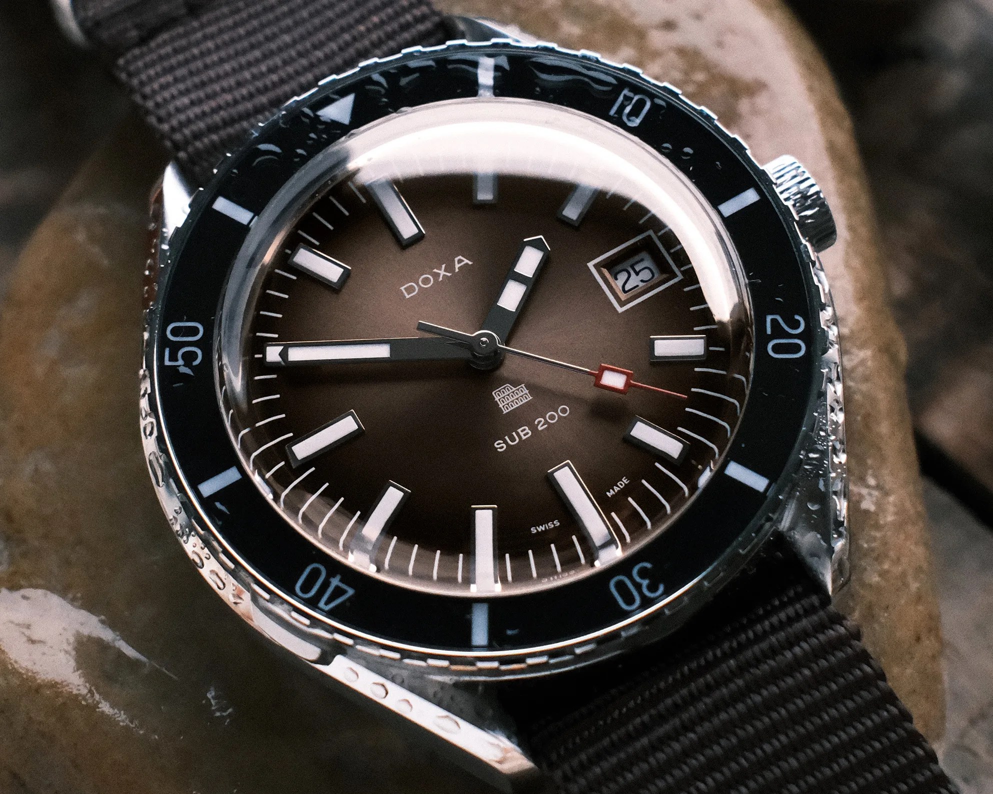 Close-up of a Doxa Sub 200 wristwatch with a brown dial, black bezel, and black fabric strap on wet stones.