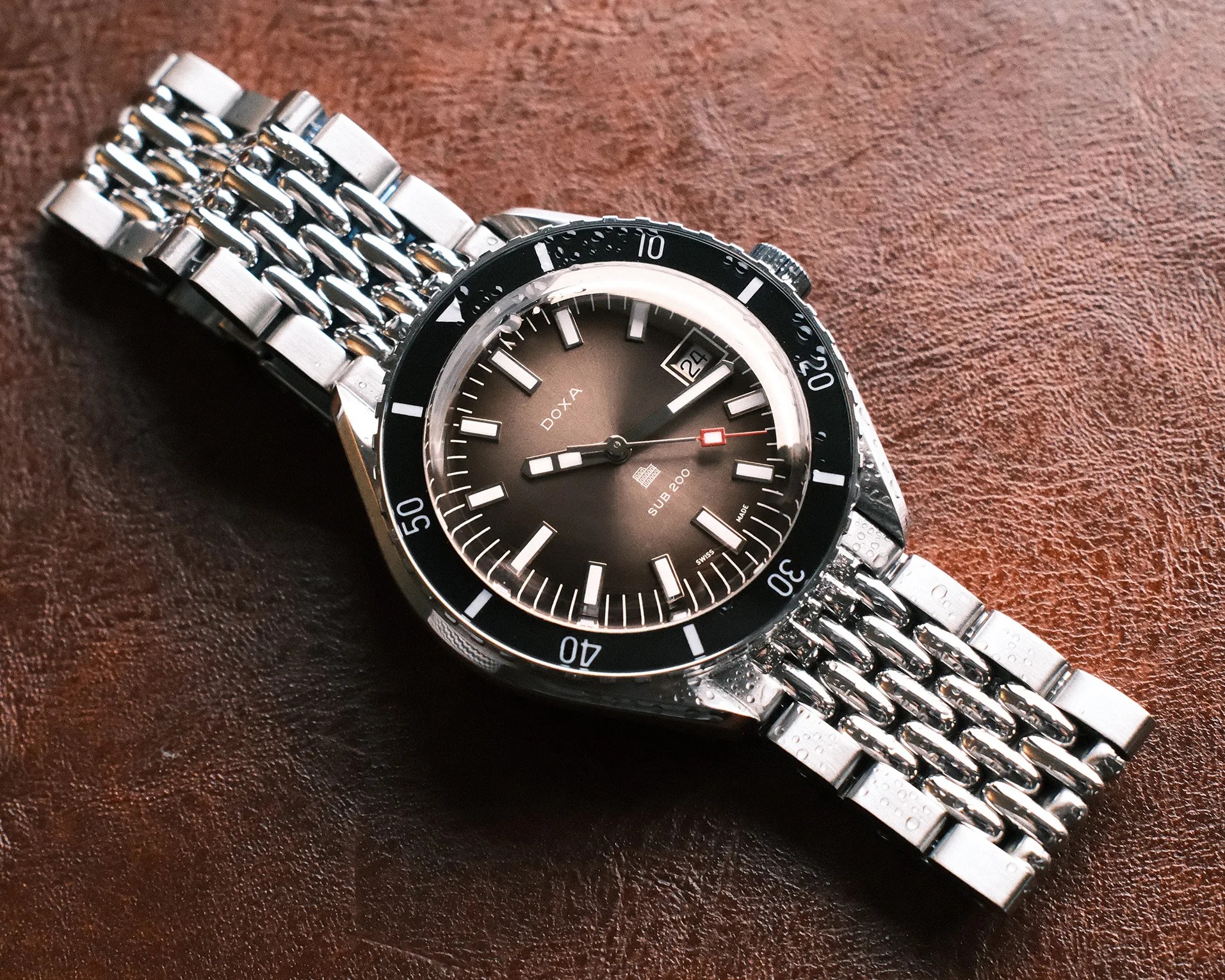Silver metal bracelet watch with black bezel and brown gradient dial showing date on a brown textured surface.