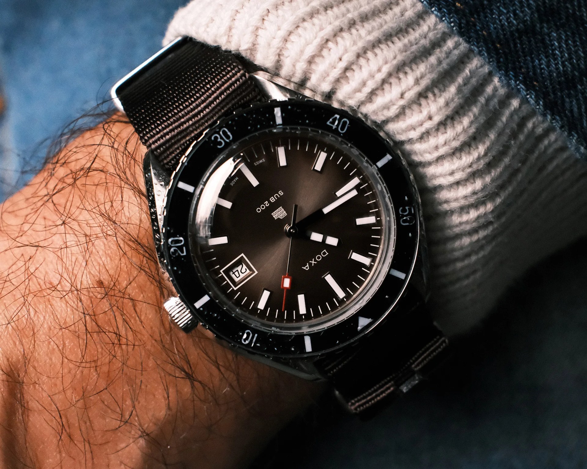 Black Doxa SUB 200 wristwatch with white markers and date window on a black fabric strap worn on a wrist.