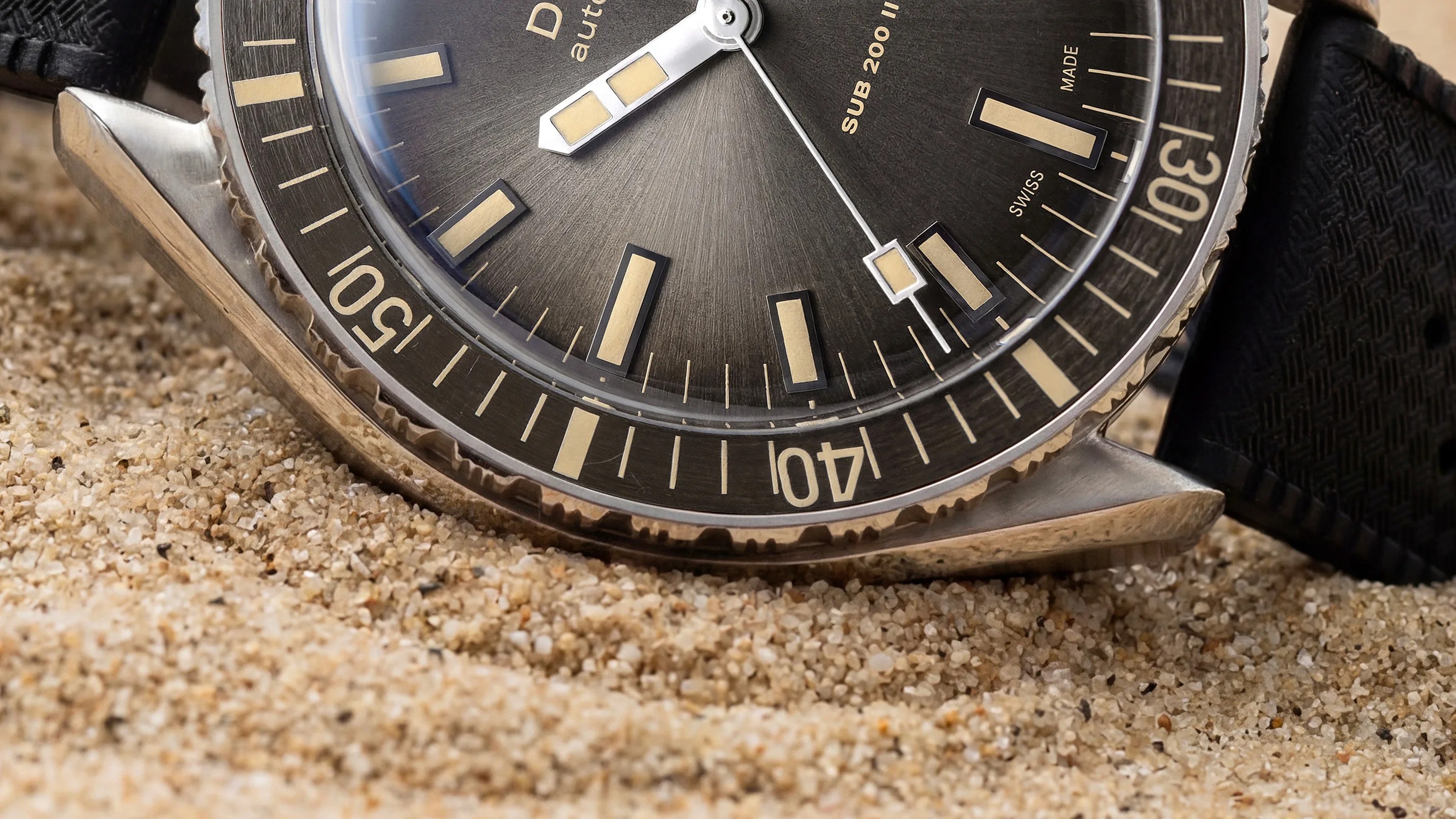Close-up of a black dive watch with beige markers and a rotating bezel resting on sand.