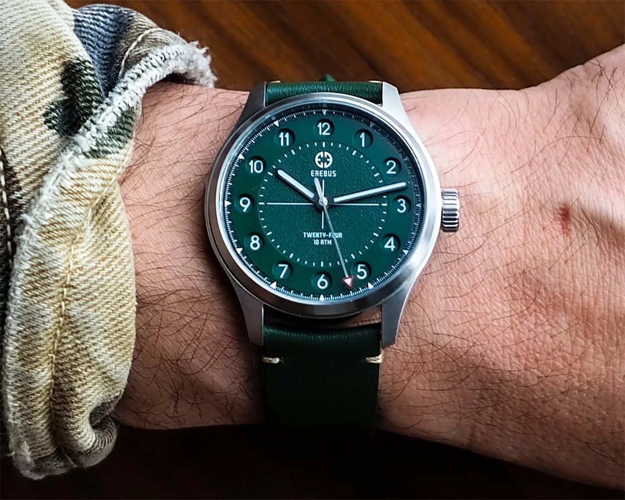 Silver wristwatch with a green dial and leather strap worn on a wrist over a plaid shirt cuff.