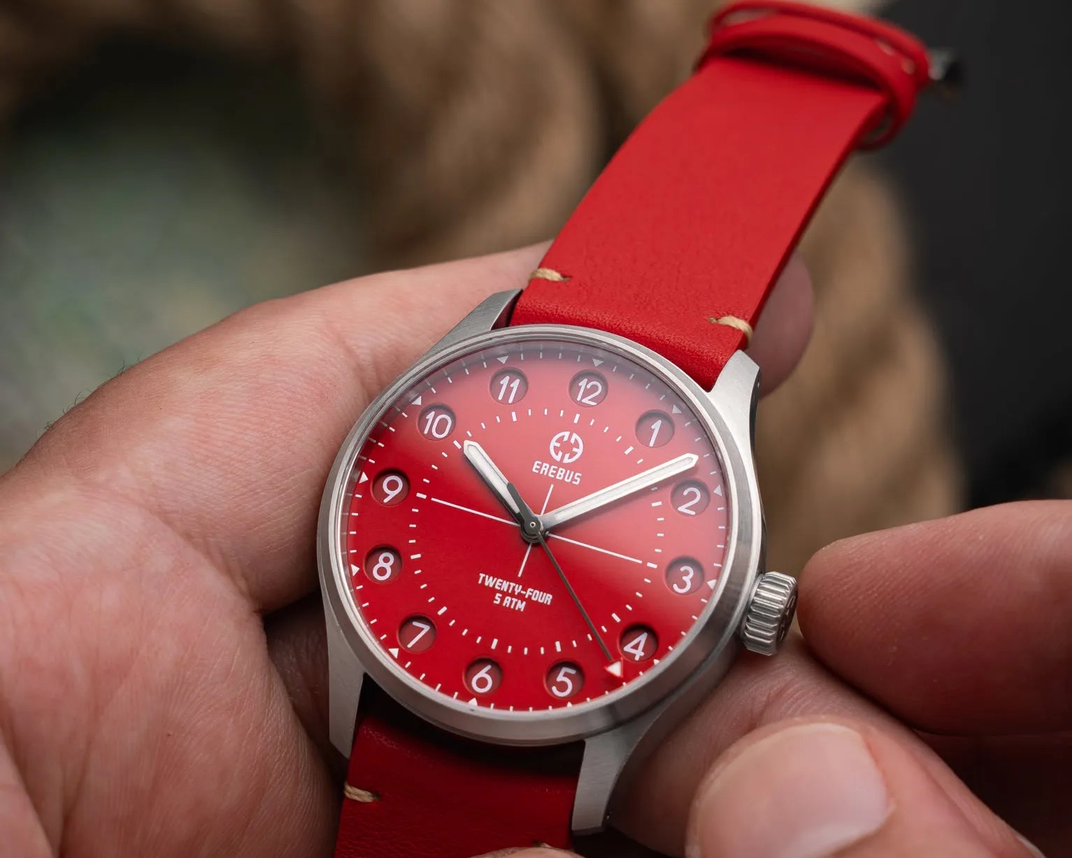 Close-up of a wristwatch with a red dial and red leather strap held in a hand, showing the brand "EREBUS" and "TWENTY-FOUR 5 ATM" on the face.