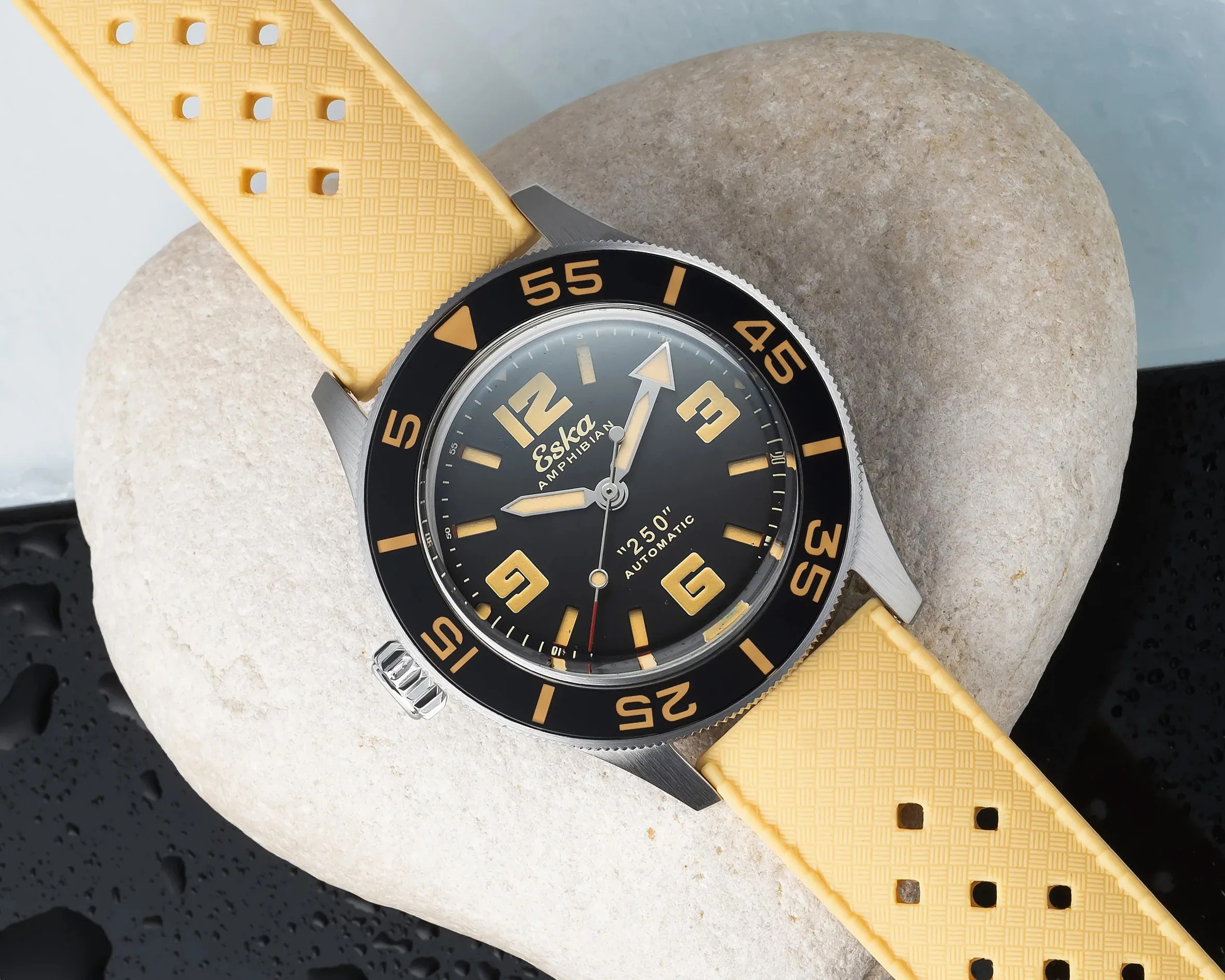 Round stainless steel dive watch with black dial, yellow numerals, and yellow perforated rubber strap on a stone.