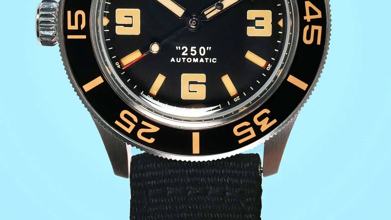 Close-up of a black watch face with beige numerals and markers, "250" AUTOMATIC text, and a black fabric strap.