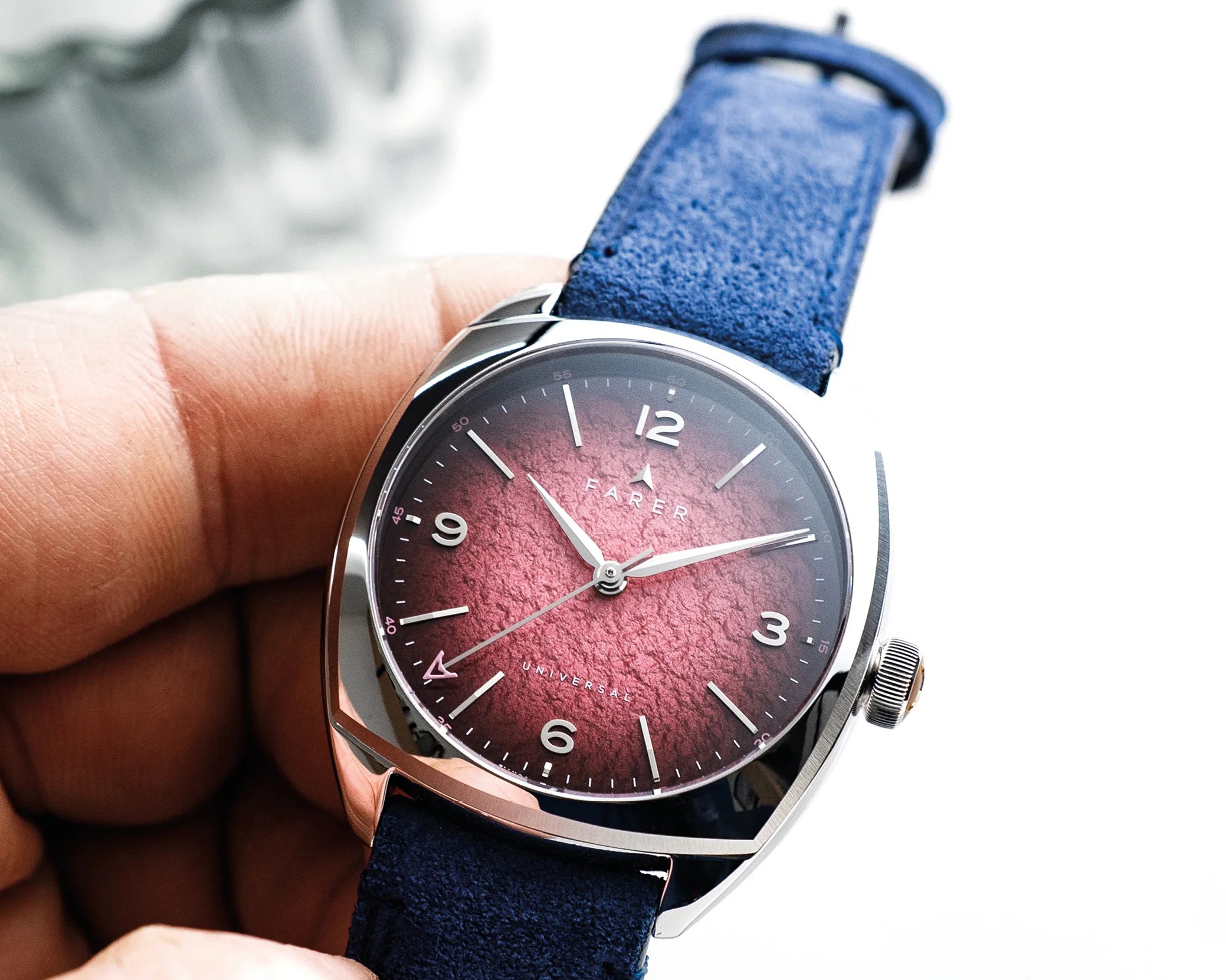Silver wristwatch with textured red dial, white numerals, and blue suede strap held in a hand.