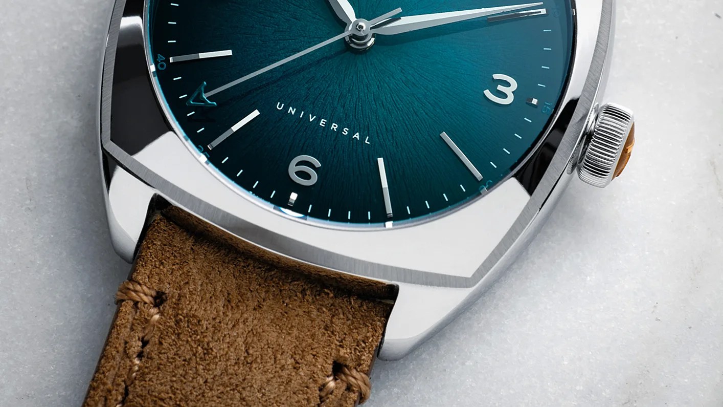 Close-up of a wristwatch with a teal textured dial, silver markers, polished steel case, and brown suede leather strap.