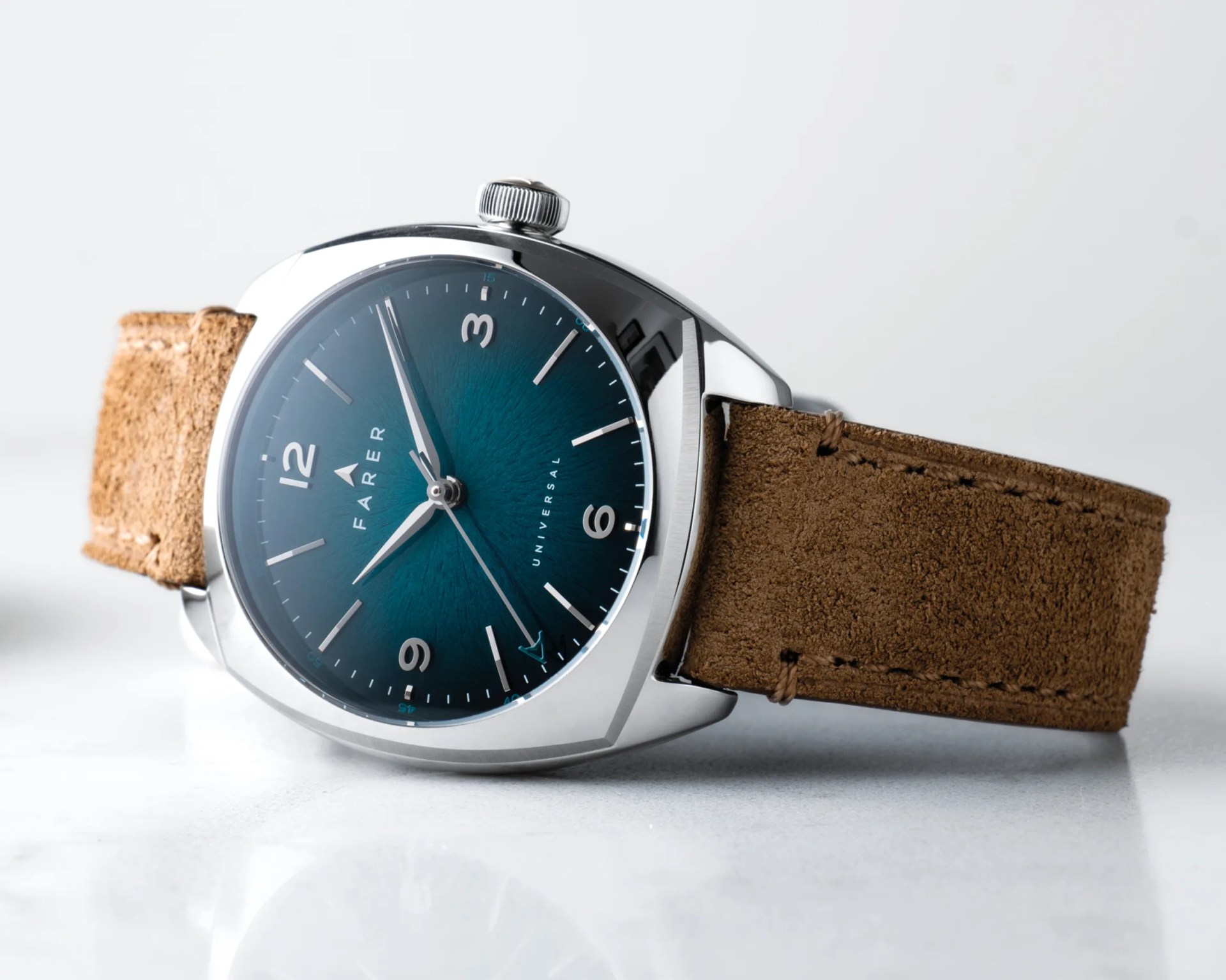 Silver wristwatch with a teal dial, white markings, and a brown suede leather strap on a white surface.